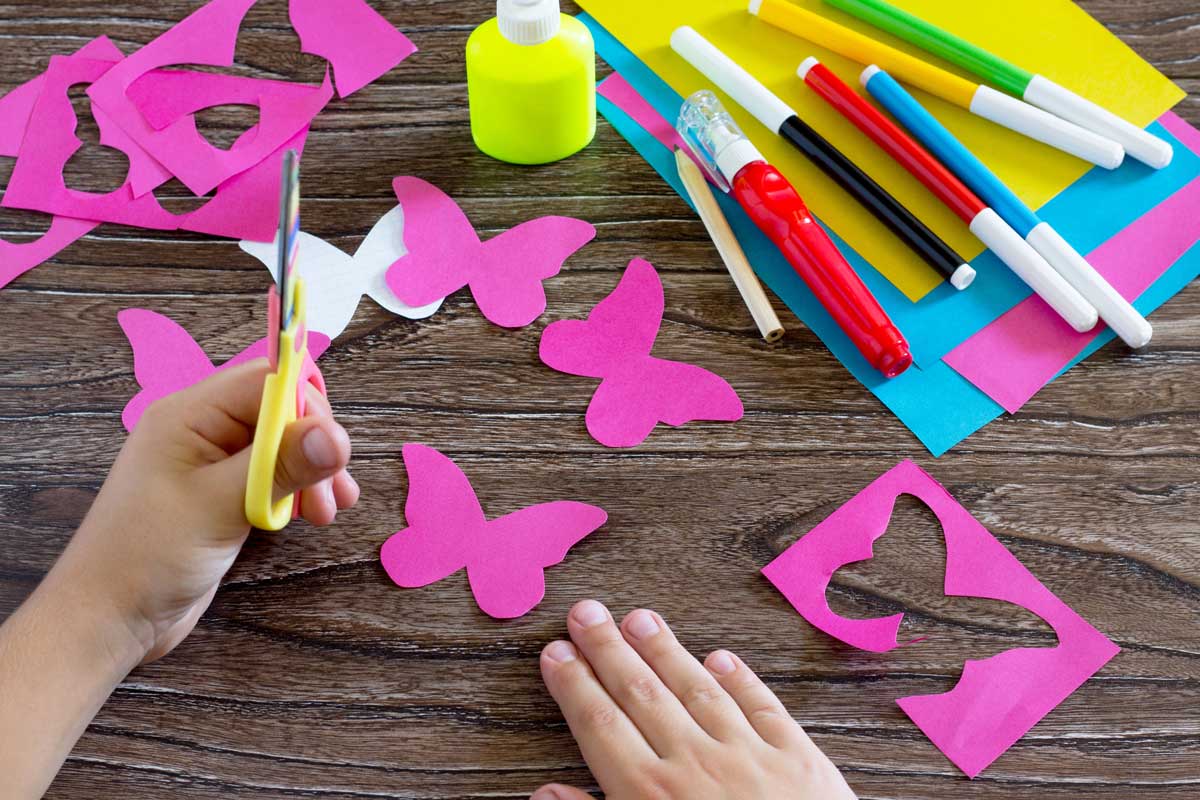 Buntes Papier, Stifte, Kleber und Kind mit Schere in der Hand, dass einen Papierschmetterling ausschneidet